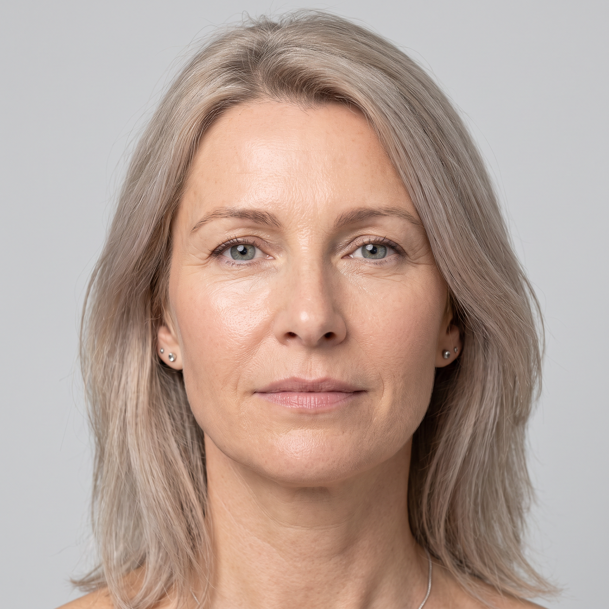 Close-up van een vrouw met grijsblond schouderlang haar en een natuurlijke, verzorgde huid, recht in de camera kijkend tegen een lichtgrijze achtergrond